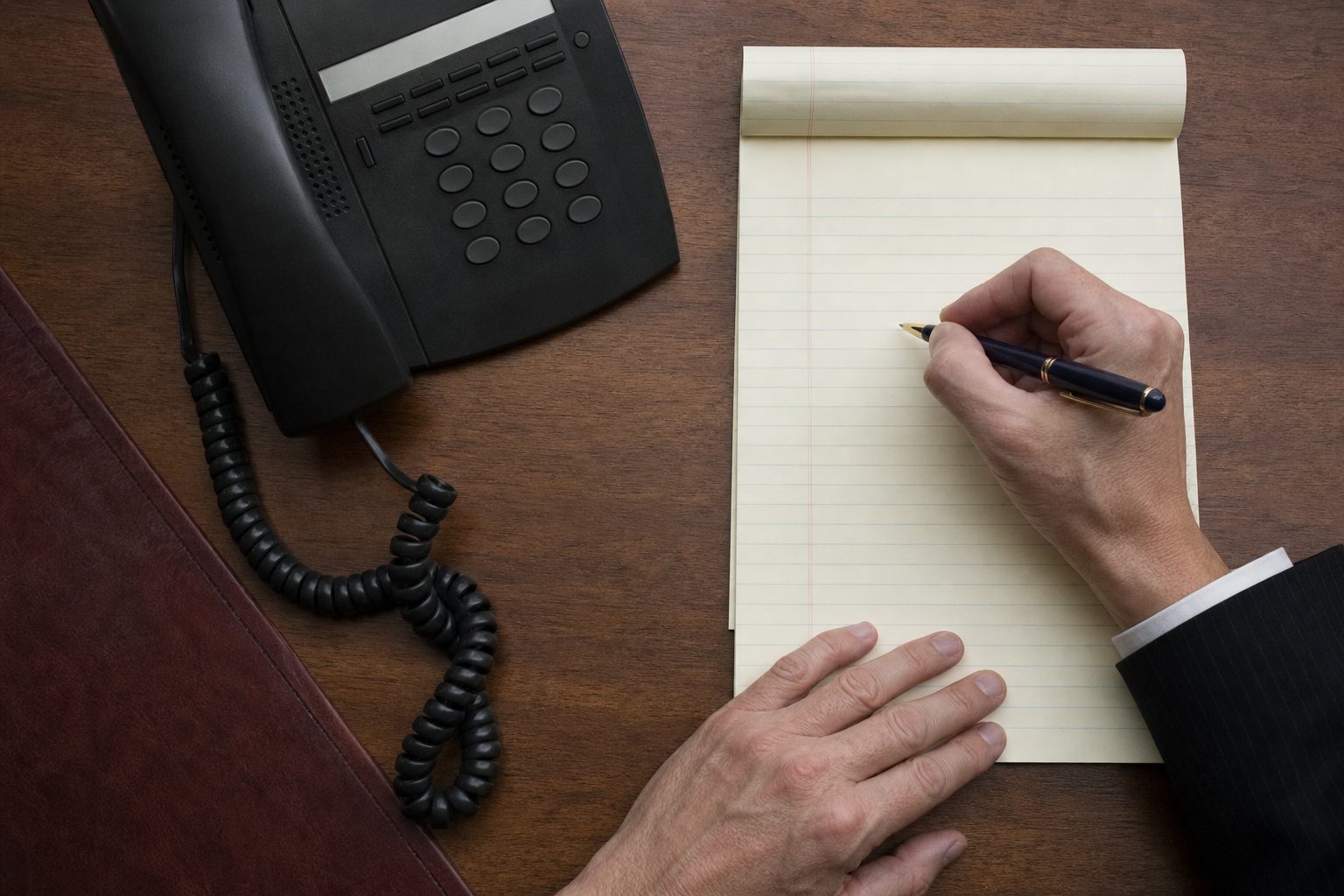 A hand writing on a ruled intake form next to a desk telephone handset in quiet daylight